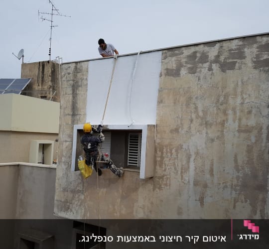 עובד בגובה צובע קיר חיצוני בעזרת חבלים